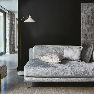 Stylowy salon z szarą sofą, poduszkami i elegancką lampą stojącą na tle ciemnej ściany. Jasne okna po lewej stronie.
