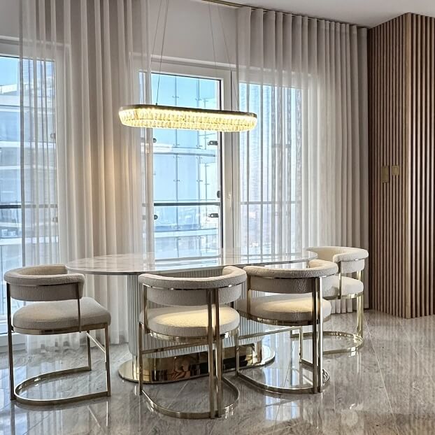 Nowoczesna jadalnia z owalnym stołem, sześcioma krzesłami i wiszącą lampą, jasne zasłony i duże okna w tle.