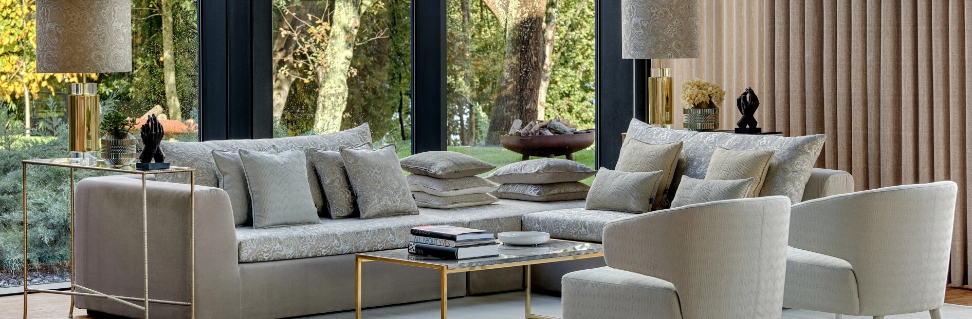 Nowoczesny salon z dużym beżowym narożnikiem, wieloma poduszkami, stolikiem i widokiem na ogród przez panoramiczne okno.