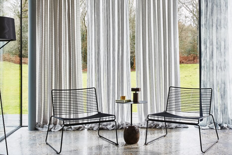 Dwa metalowe krzesła i stolik przy wysokich oknach z zasłonami, z widokiem na trawnik i drzewa.