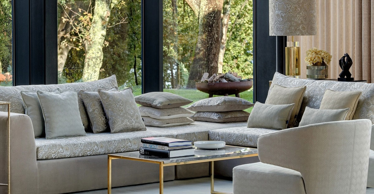 Nowoczesny salon z szarą kanapą, stosem poduszek, stolikiem kawowym i dużymi oknami z widokiem na ogród.