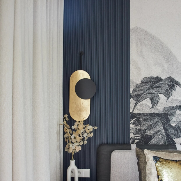 Dekoracyjna lampa ścienna na ciemnym tle, w pobliżu białej zasłony i grafiki z liśćmi; obok wazon z suszonymi kwiatami.