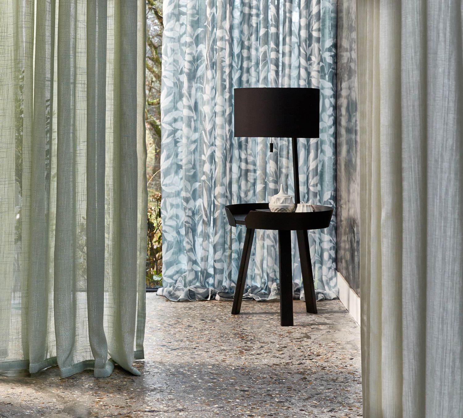 Lampa na stoliku przy zasłonach w odcieniach zieleni i niebieskiego z roślinnym wzorem, stojących na kamiennej podłodze.