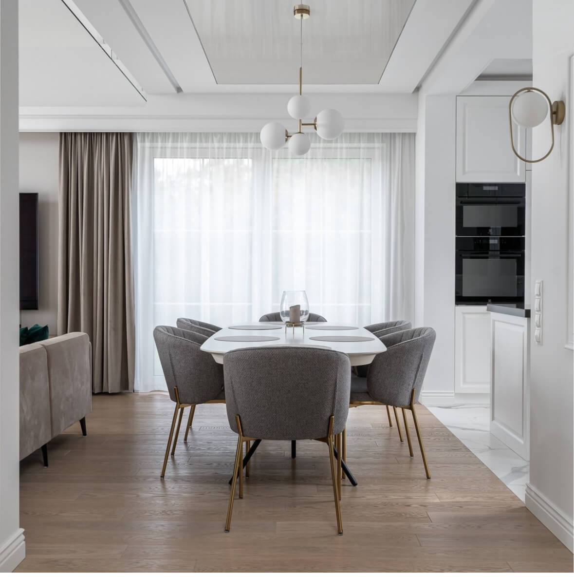 Elegancka jadalnia z białym stołem, szarymi krzesłami i nowoczesnym żyrandolem, oświetlona naturalnym światłem z dużego okna.