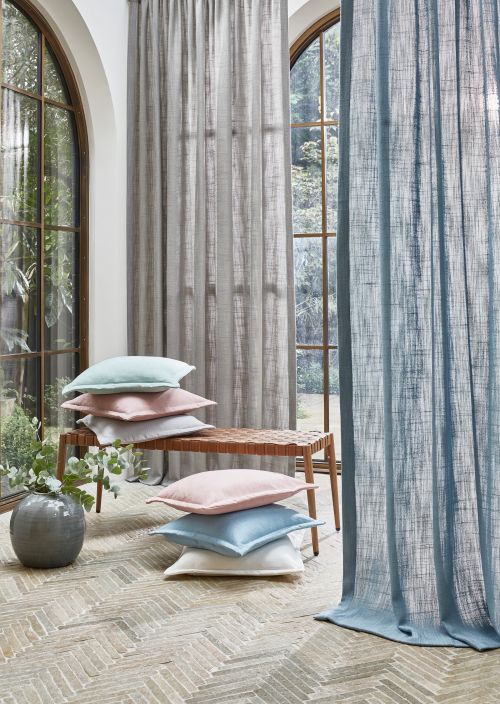 Stosy pastelowych poduszek na ławce przy dużych oknach z zasłonami, w jasnym, eleganckim wnętrzu.