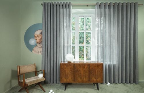 Okno z szarymi zasłonami, zielone ściany, plakat z bańką, drewniany fotel i komoda, białe kule na podłodze.