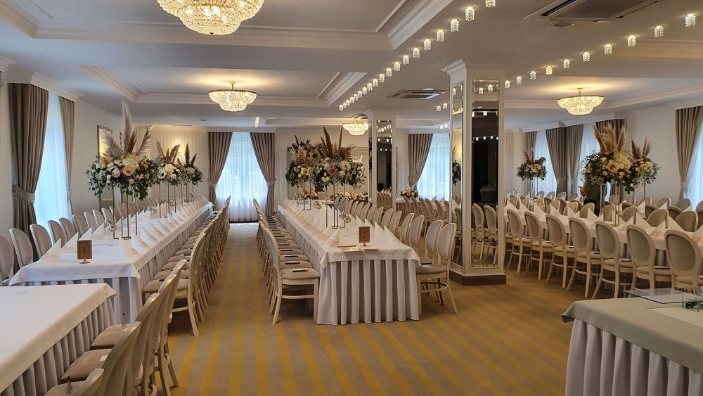 Elegancka sala bankietowa z długimi stołami, ozdobiona kwiatami i wiszącymi żyrandolami.