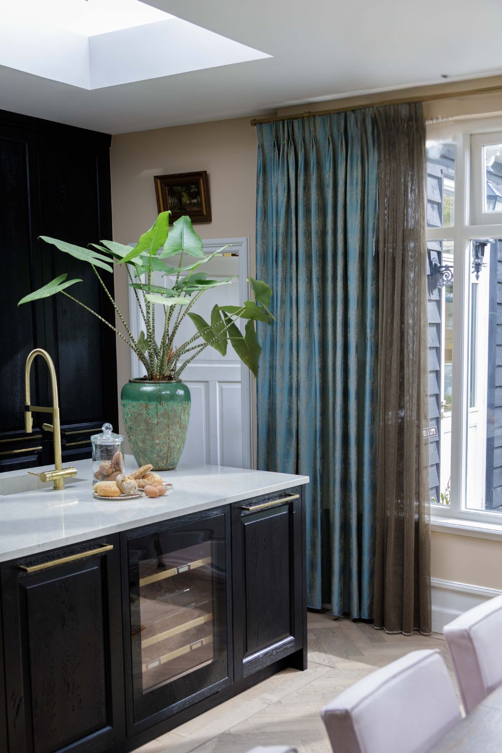 Kuchnia z ciemnymi szafkami, rośliną na blacie, złotym kranem i niebieskimi zasłonami przy jasnym oknie.