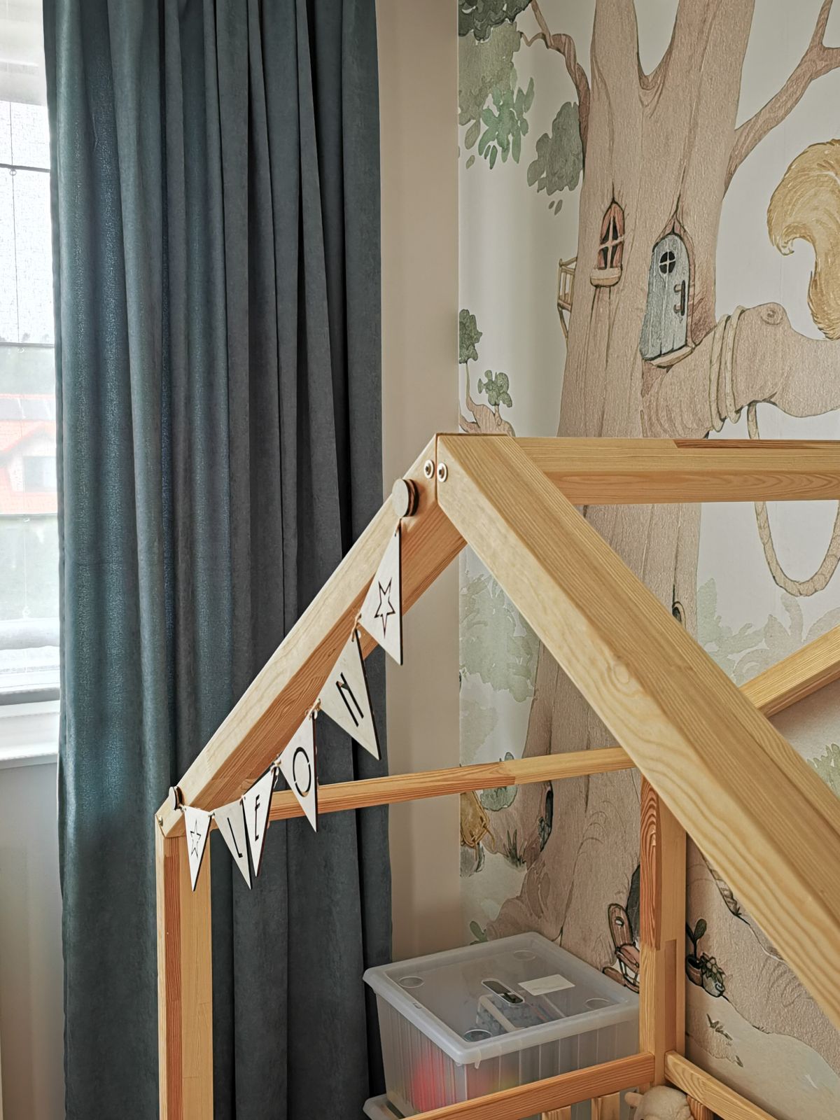 Drewniana rama łóżka w kształcie domku z flagową girlandą, na tle ozdobnej ściany z motywem drzewa i niebieskich zasłon.