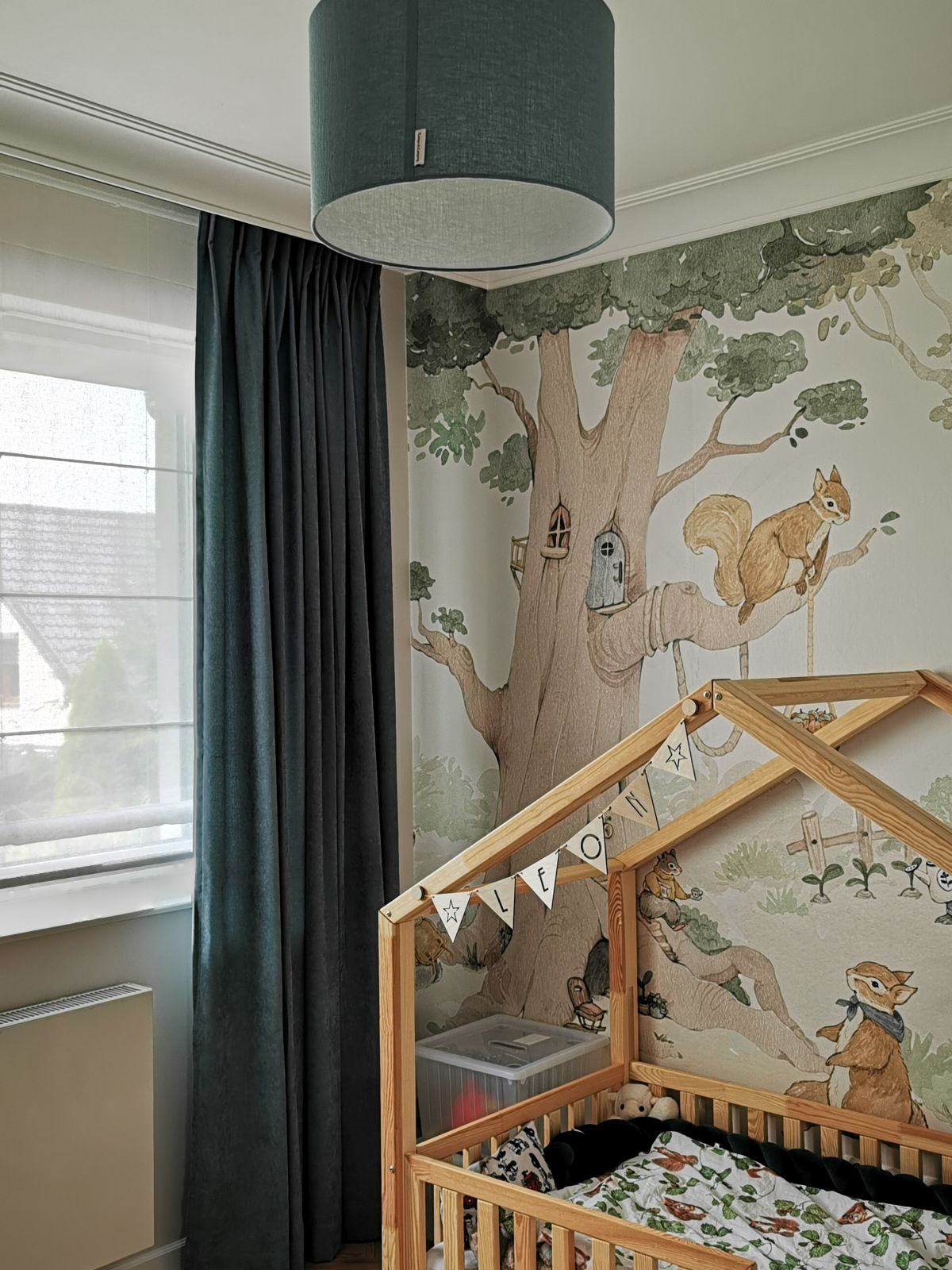 Dziecięcy pokój z łóżeczkiem w kształcie domku, las na ścianie, zasłony i lampa sufitu w ciemnym kolorze.
