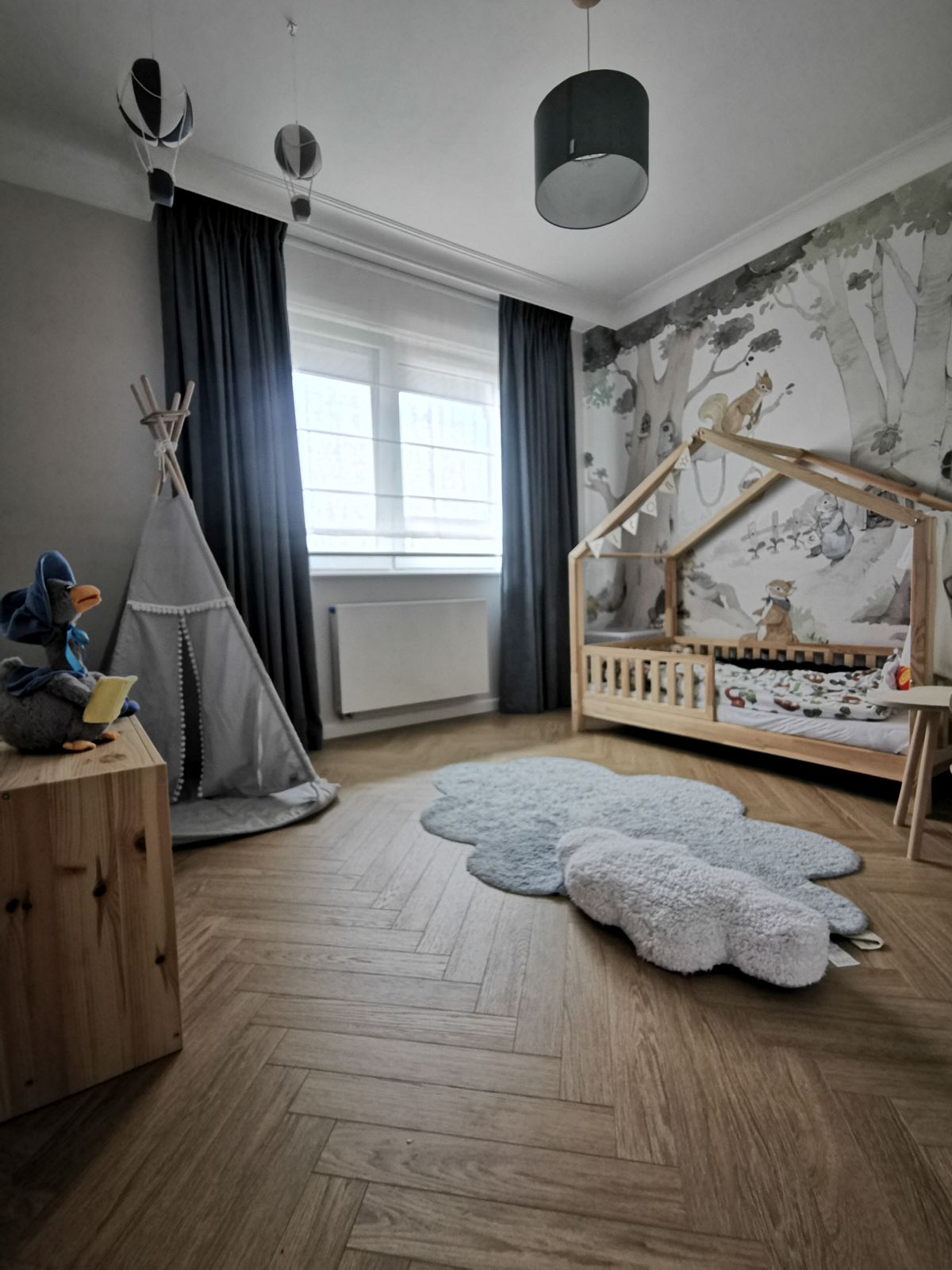 Pokój dziecięcy z łóżeczkiem, tipi, dywanikiem w kształcie chmurki i leśną tapetą na ścianie.