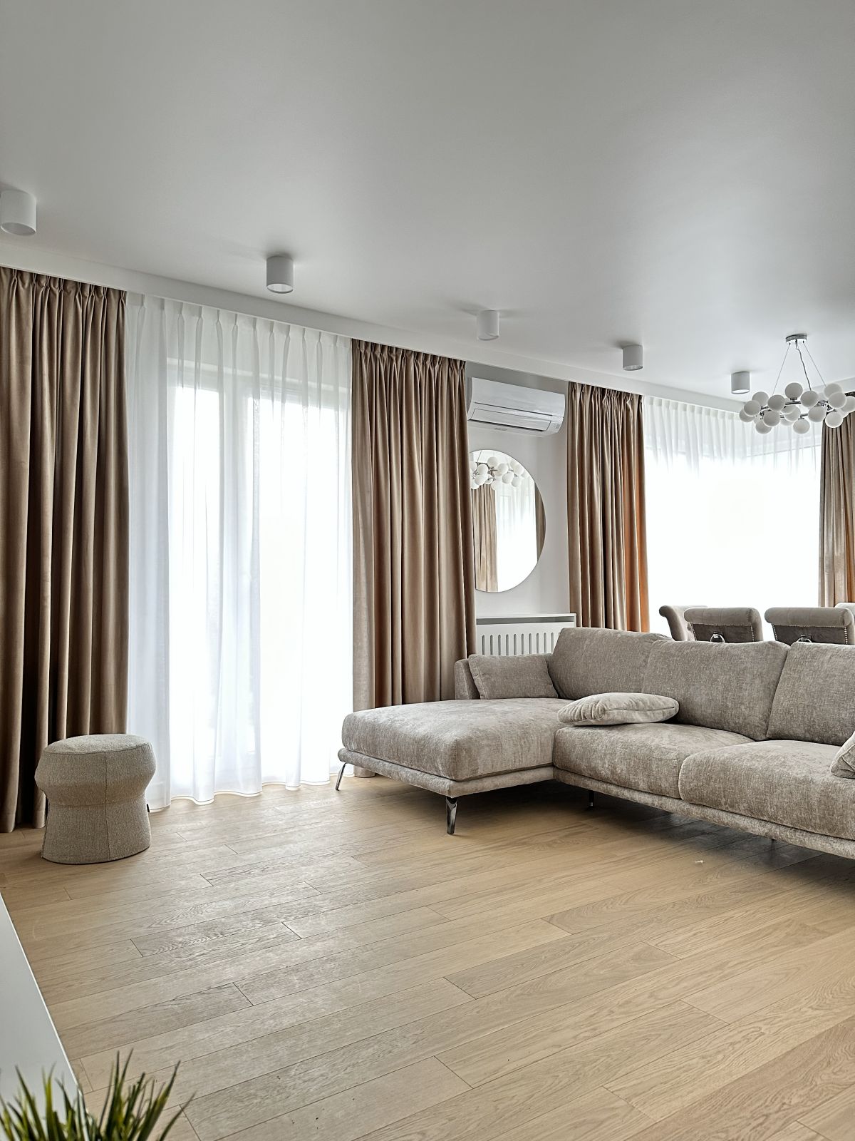 Nowoczesny salon z beżową sofą, drewnianą podłogą, jasnymi zasłonami i okrągłym lustrem na ścianie.