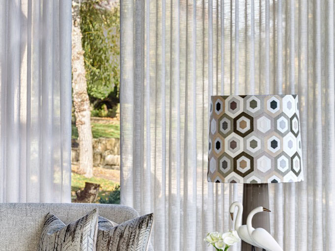 Pokój z jasnymi zasłonami, lampą o geometrycznym wzorze i dekoracyjnymi poduszkami, widok na ogród przez okno.